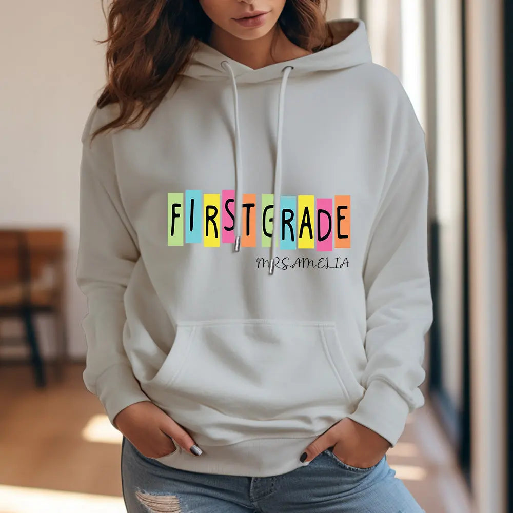 Lifestyle shot of teacher in classroom wearing personalised first grade hooded sweatshirt while holding books — practical polyester teacher customised hoodie.