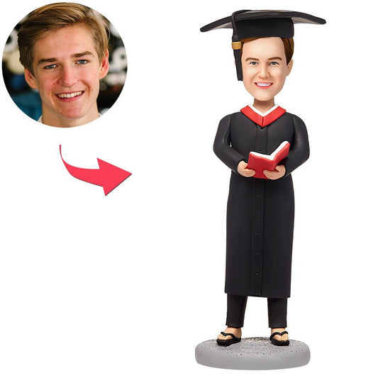 Personalised male graduate bobblehead in mortarboard and gown holding an engraved red book — custom polymer clay head modelled from your photo; perfect personalised graduation gift for him in the UK and Australia