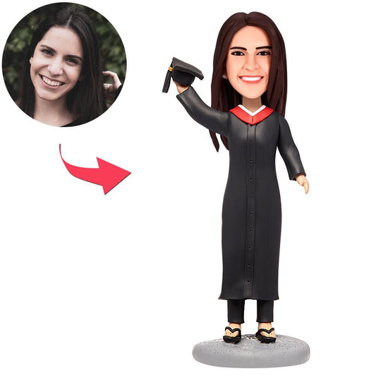 Personalised female graduate bobblehead in black gown holding a bachelor hat, handcrafted polymer-clay figurine with a customised head and engraved text — custom graduation gift and keepsake for her